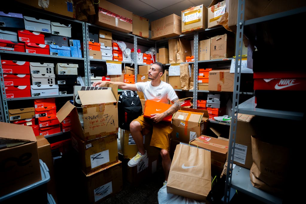 A man organizing various shoe boxes in a cluttered store room filled with shelves and cartons.