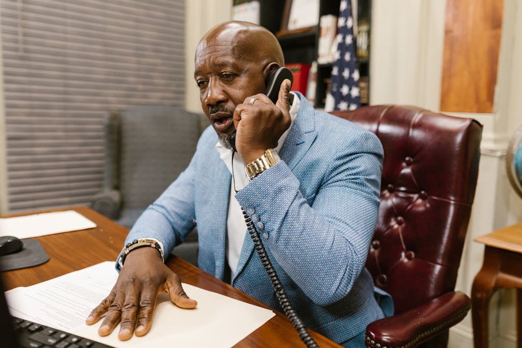 African American man in a blue suit speaking on the phone in a professional office setting.