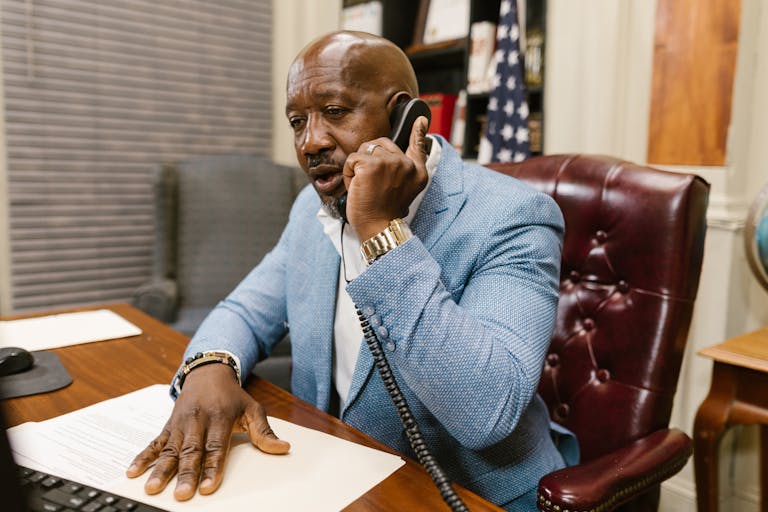 African American man in a blue suit speaking on the phone in a professional office setting.