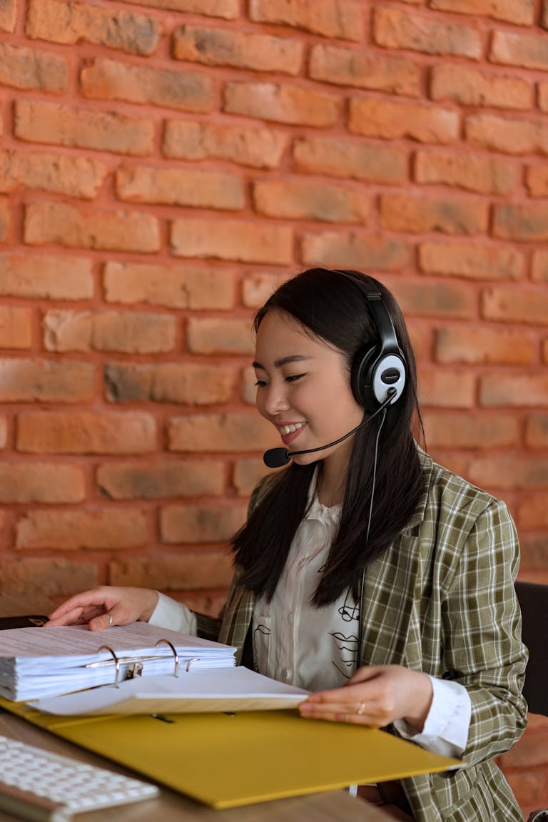 Professional Asian woman wearing a headset, working efficiently at an office with a binder, smiling and focused.